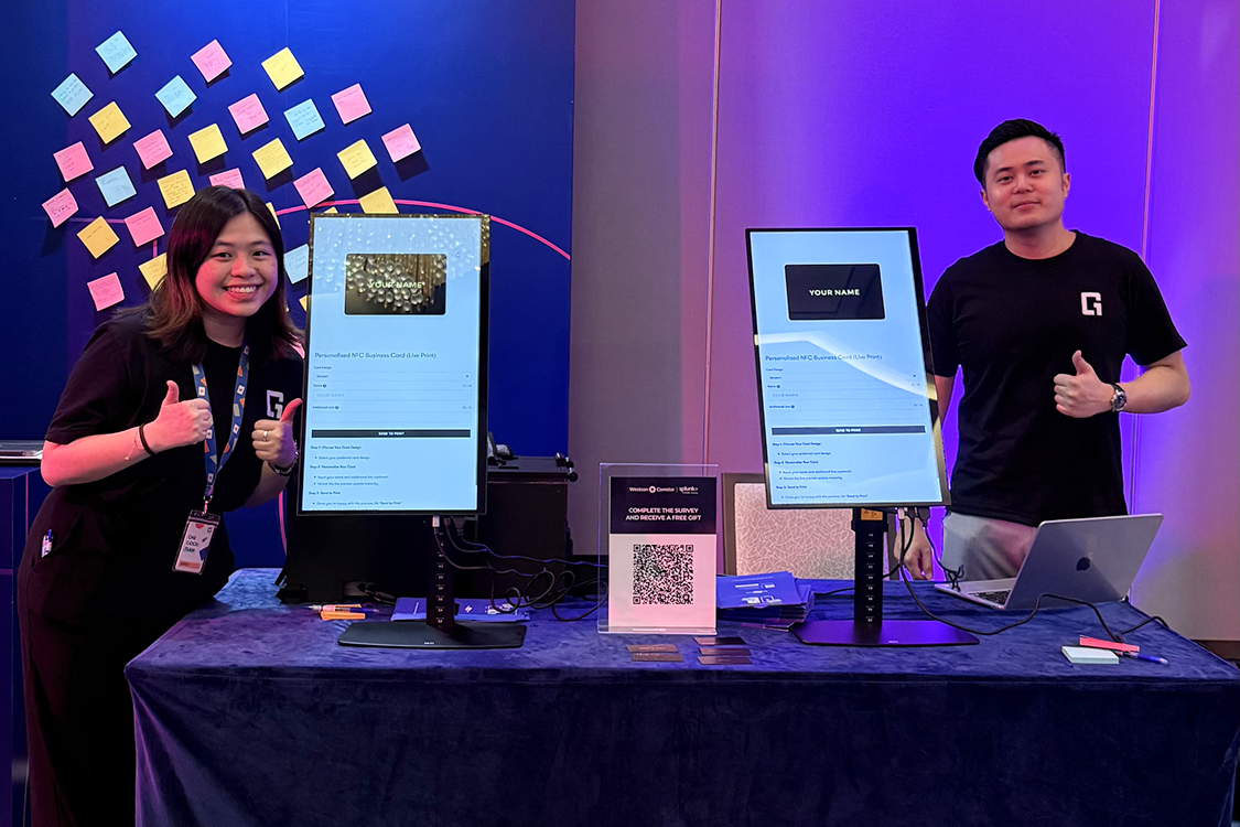 Two team members giving a thumbs up beside live card-printing kiosks at an event booth